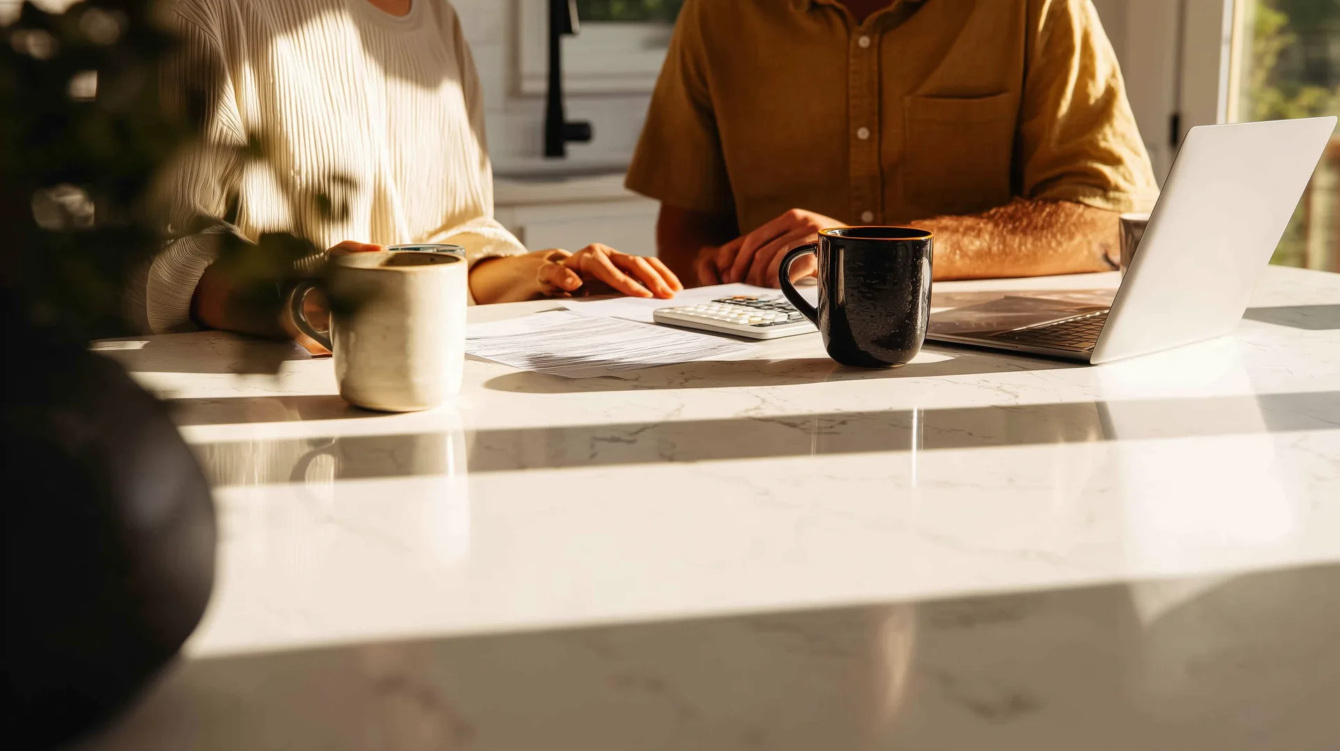 Two people reviewing documents at a kitchen island with laptop and calculator in morning light